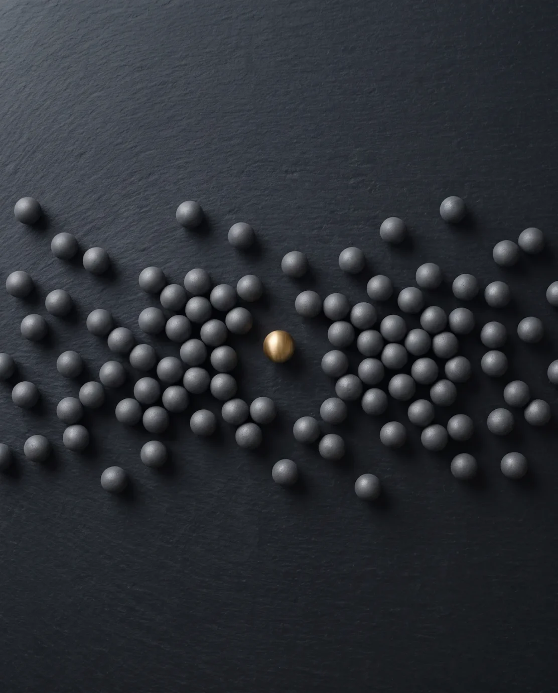 Top-down view of scattered matte grey spheres on a dark surface, one of them in brushed gold.
